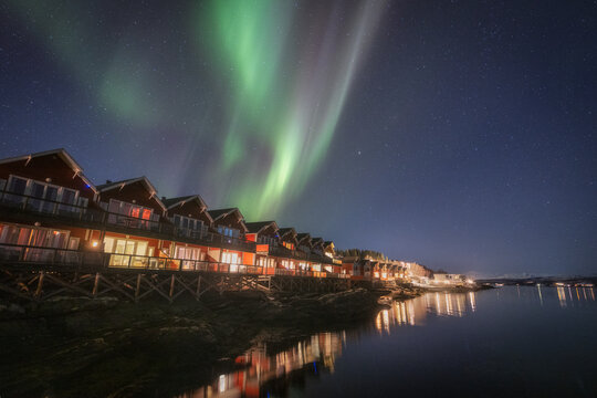 View of the aurora borealis dances above a row of warmly lit cabins nestled along the tranquil waters edge, Troms, Troms, Norway.