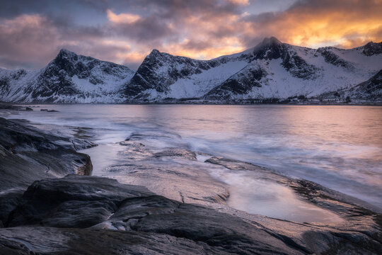 View of twilight hues painting the sky above snow-dusted peaks reflected in the tranquil waters, kissed by the rocky shoreline, Troms&Atilde;&cedil;, Troms, Norway.