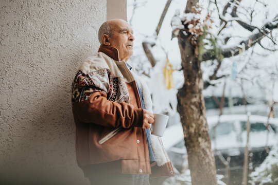 Senior man enjoying a cup of coffee outside during winter, dressed warmly and gazing outward, surrounded by snowy scenery, evoking calmness and thoughtfulness.