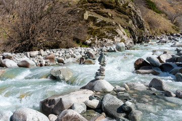 Ala-Archa Park, Fast flowing river with balanced rock cairn, rugged boulders, mossy hillside, bare trees, clear water, and untouched landscape ideal for mountaineering and backcountry exploration
