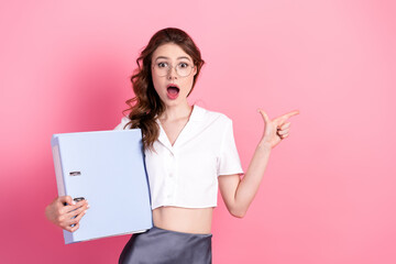 Young fashionable woman with glasses pointing in surprise, holding a blue folder, against a vibrant...