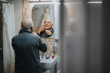 Senior man maintaining personal grooming by shaving in front of a bathroom mirror, expressing...