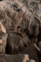
A woman with voluminous dark curly hair in a white bikini and large hoop earrings reclines on sunlit rocks, holding a vintage camera, sitting at Rocky beach Marbella Spain