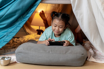 A cheerful young girl lies under a table, enjoying a mobile game on her smartphone, capturing childhood joy and screen time fun.