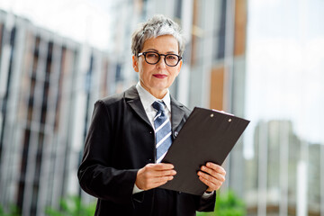Confident mature businesswoman holding clipboard outdoors with modern buildings in urban setting,...