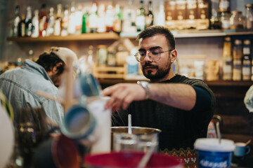 A man with glasses, focusing on making drinks at a cozy bar counter with bottles in the background. The interior design features warm lighting and modern elements.