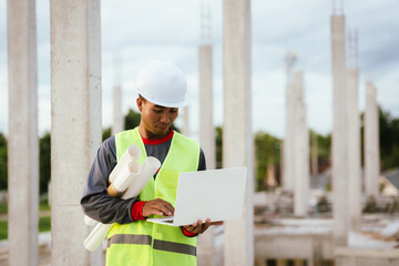 A construction worker wearing a yellow helmet and safety vest examines blueprints on a building...