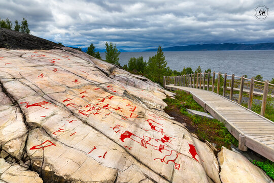 View of ancient red rock carvings etched into a sloping stone surface, meeting the tranquil blue waters beyond a wooden pathway, Alta, Finnmark, Norway.