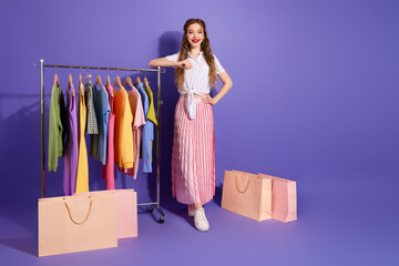 Fashionable young woman in stylish outfit posing with colorful clothes rack against purple...