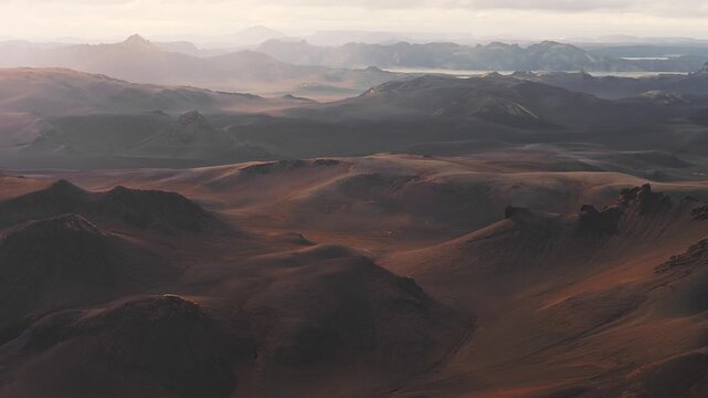 Sweeping aerial pan over Iceland's rugged volcanic terrain, featuring craters, ridges, distant mountains, and glimmering lakes in golden sunlight.