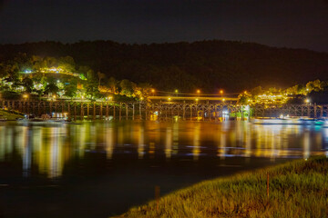 Fototapeta premium The enchanting night view of Wooden Mon Bridge at Sangkla Buri Kanchanaburi, Thailand, this cityscape reflects city lights on the water with a bridge silhouetted against the evening sky.