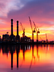 Naklejka premium Industrial cranes and chimneys silhouetted against a vibrant sunset sky, reflected in calm water at a construction site or refinery.