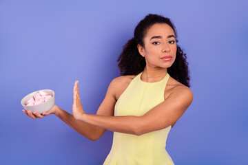 Young woman in yellow dress refusing bowl of pink marshmallows against purple background,...