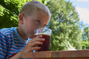 a boy drinks cold kvass in the park, a boy sits at a wooden table in a summer cafe and drinks kvass with foam