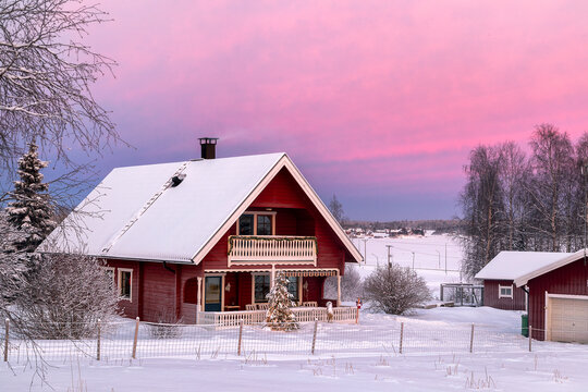Haparanda, Sweden - 16 July 2025: View of a red wooden house with a snow-covered roof under a vibrant pink and purple sky, framed by frosted trees in a winter wonderland.