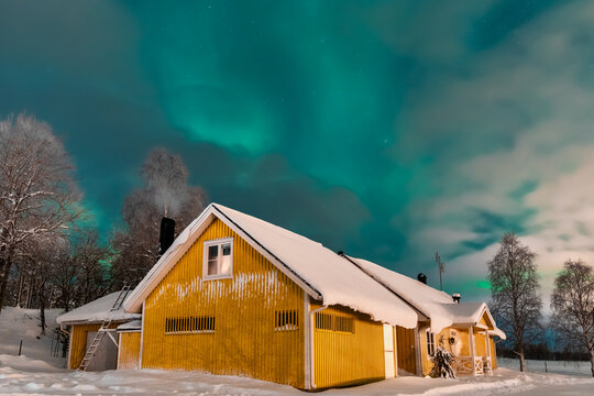 Haparanda, Sweden - 16 July 2025: View of a vibrant yellow house glows warmly beneath the ethereal dance of the Northern Lights, a stark contrast to the snow-laden landscape.