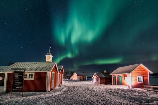 Haparanda, Sweden - 16 July 2025: View of the aurora borealis dancing vibrantly above the snow-covered landscape and rustic red buildings.