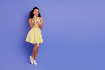 Chic stylish woman holding a smartphone, dressed in a trendy yellow dress, standing against a solid...