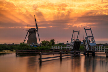 Kinderdijk, Netherlands - 16 July 2025: View of a historic windmill standing tall against a fiery sunset, its reflection shimmering in the tranquil canal waters.