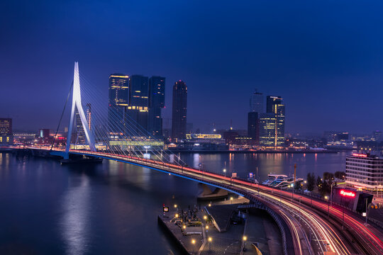 Fototapeta Rotterdam, Netherlands - 16 July 2025: View of the Erasmus Bridge illuminated against the night sky, with the city's modern architecture reflecting in the dark waters of the Nieuwe Maas.