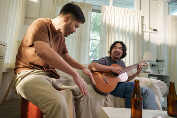 Two Asian men taking turns strumming acoustic guitar, enjoying drinks and conversation in a relaxed...