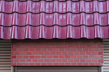 Detail of a building facade with a shiny purple tiled roof, red brick wall, and closed shutter.