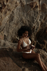 Naklejka premium A young woman with curly hair smiles, holding a vintage camera while sitting on sunlit rocks by the sea.