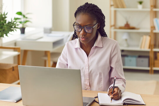 Portrait of a concentrated serious african american business woman sitting in a shirt and glasses at the desk on her workplace and working on laptop at office looking at the monitor screen. - Powered by Adobe
