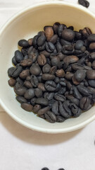 A bowl of freshly roasted coffee beans is dark in color, on a white background.