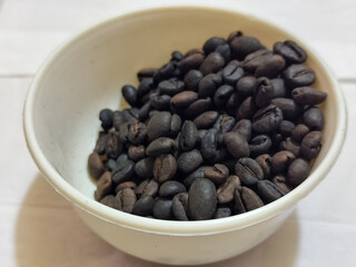 A bowl of freshly roasted coffee beans is dark in color, on a white background.