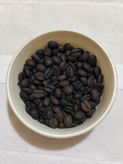 A bowl of freshly roasted coffee beans is dark in color, on a white background.
