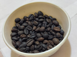 A bowl of freshly roasted coffee beans is dark in color, on a white background.