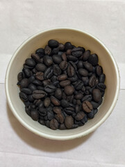 A bowl of freshly roasted coffee beans is dark in color, on a white background.