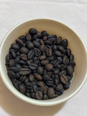 A bowl of freshly roasted coffee beans is dark in color, on a white background.