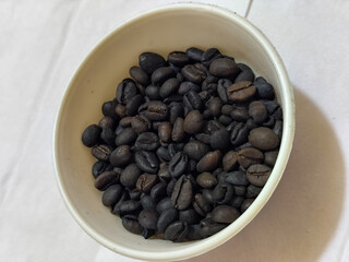 A bowl of freshly roasted coffee beans is dark in color, on a white background.
