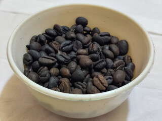 A bowl of freshly roasted coffee beans is dark in color, on a white background.
