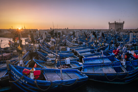 Essaouira, Morocco - 16 July 2025: View of the sun casting golden light over the bustling port, illuminating the blue fishing boats and the ancient Skala de la Kasbah fortress.
