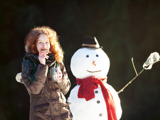 Pretty red-haired girl drinking tea and eating gingerbread outdoors with snowman, sunny winter day