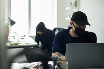 Young Caucasian male hacker wearing cap and mask using laptop at desk in modern office,...