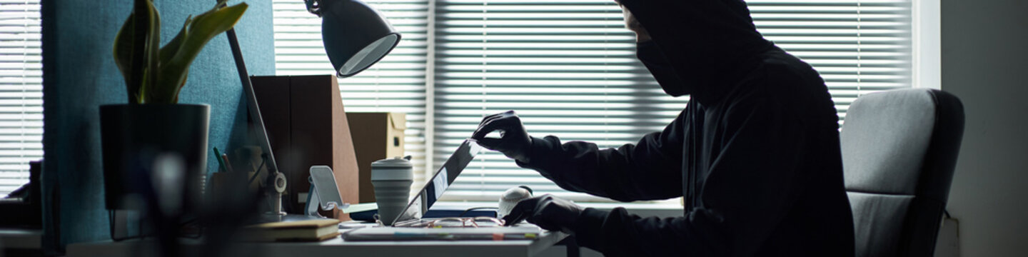 Header of Caucasian young man wearing black hoodie and gloves sitting at desk hacking into corporate laptop in dark office environment, blinds closed, engaging in secretive computer activity