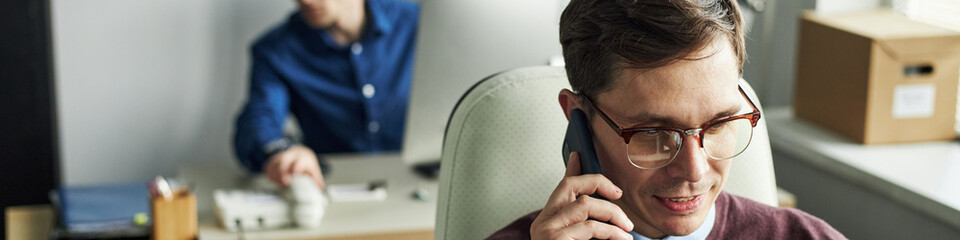 Header of Caucasian man wearing glasses talking on phone in office foreground, young male colleague...