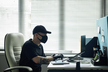 Male hacker wearing black mask and gloves sitting at desk typing on computer with tablet and...
