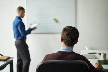 Fototapeta premium Caucasian young man sitting at desk listening to colleague standing and presenting information on whiteboard during business meeting in corporate office