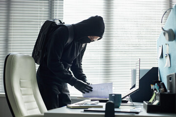 Young Caucasian man wearing black hoodie and gloves searching and stealing through documents on...