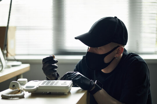 Man wearing black cap and mask tampering with landline phone using screwdriver in corporate office, planting spy bug for eavesdropping on business conversations, engaging in suspicious activity