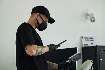 Young Caucasian man wearing black mask and gloves standing in modern office holding smartphone and...