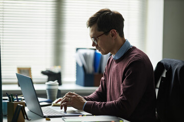 Young man working on laptop at desk in office, wearing eyeglasses, focusing on screen, typing on...