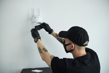 Young male robber wearing black gloves and mask covering security camera with duct tape to disable,...