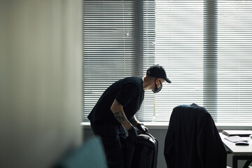 Caucasian man wearing black cap and face mask bending over desk while holding bag in office,...