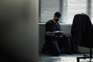 Caucasian young male spy wearing black mask and cap kneeling on floor reading business documents...